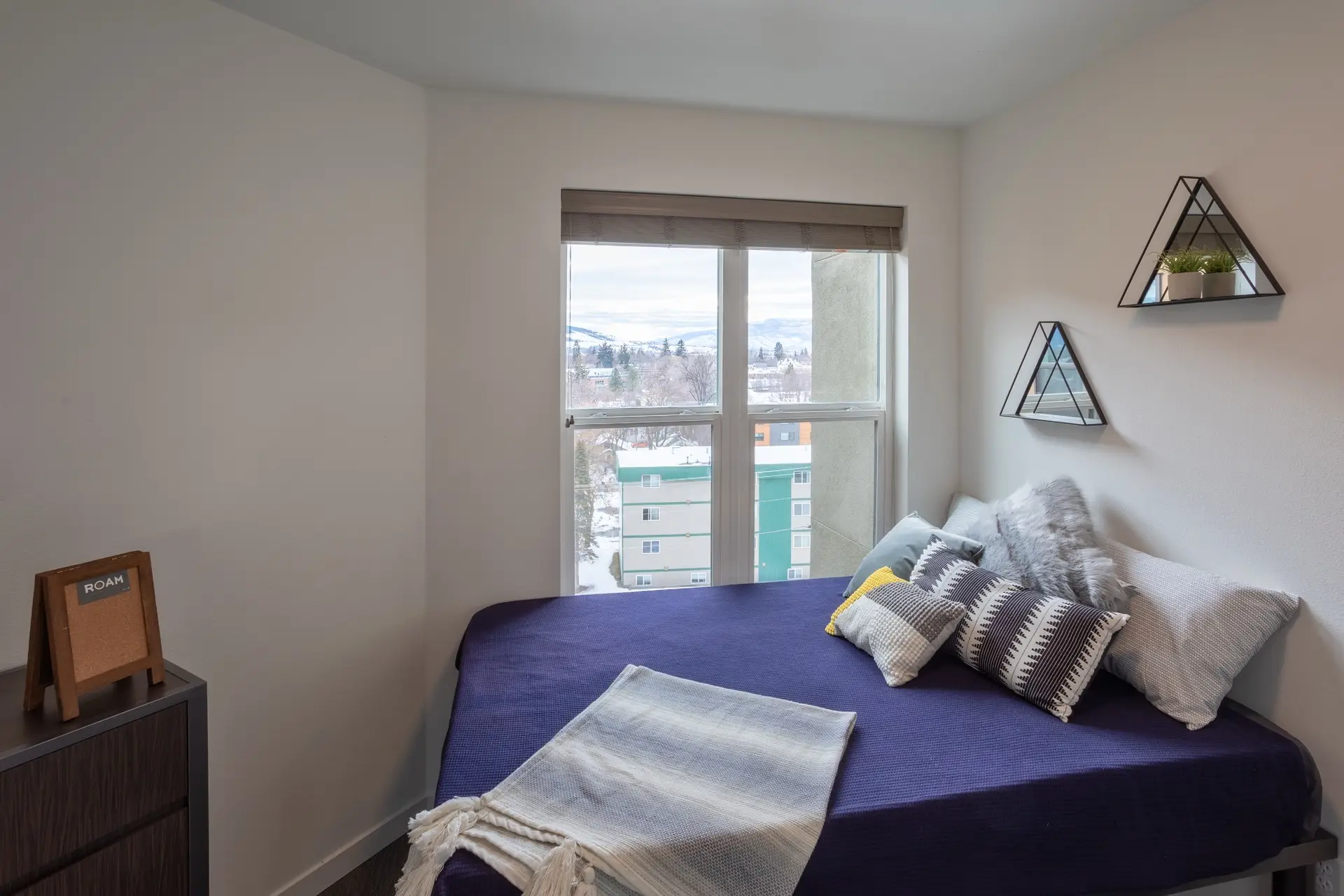 A bed made with blue linens and several decorative pillows next to a large window with a view of snowy mountains outside.