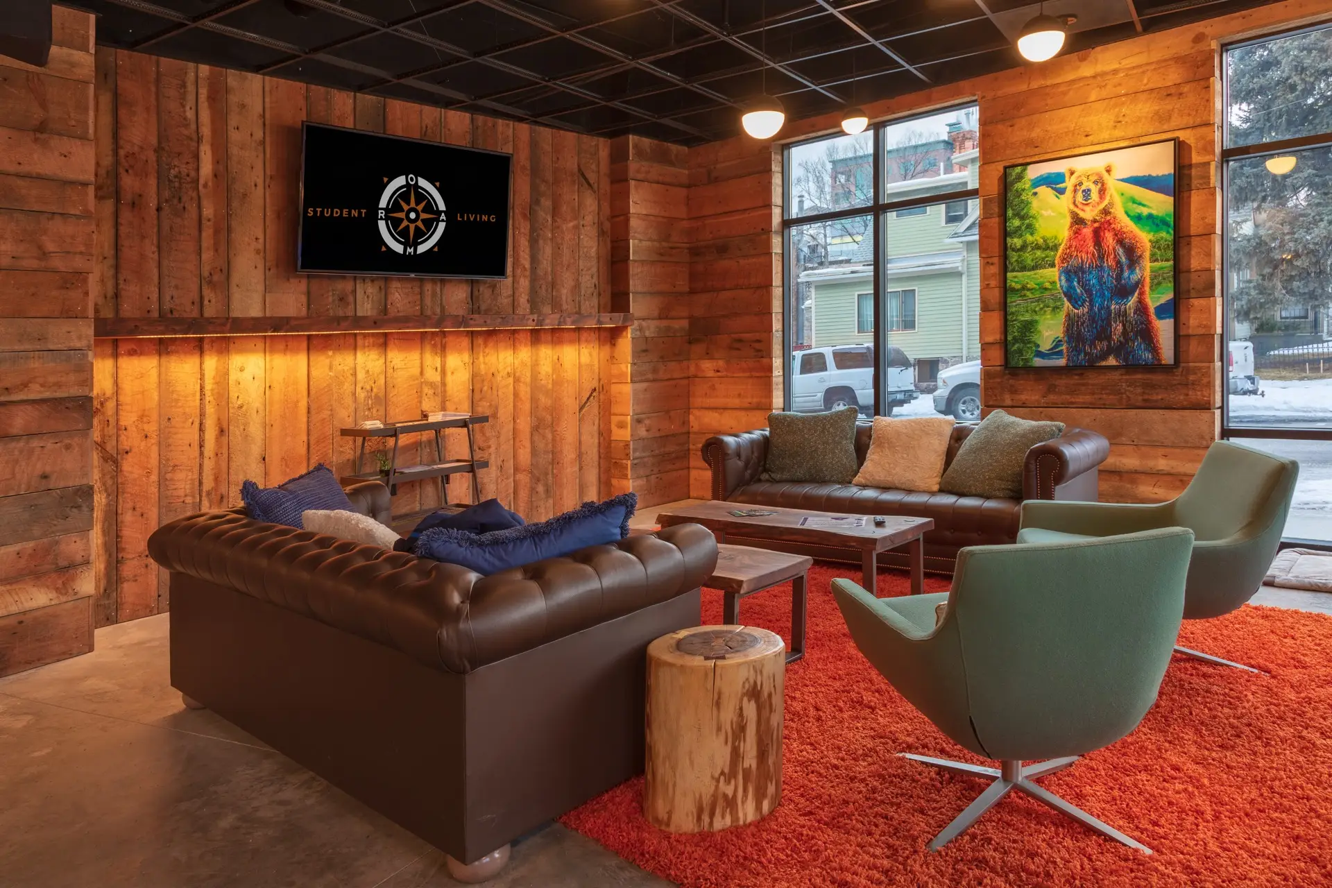 Community Area with an orange rug, rustic wood paneling on the walls, two large leather sofas, two modern green arm chairs, and rustic coffee tables.