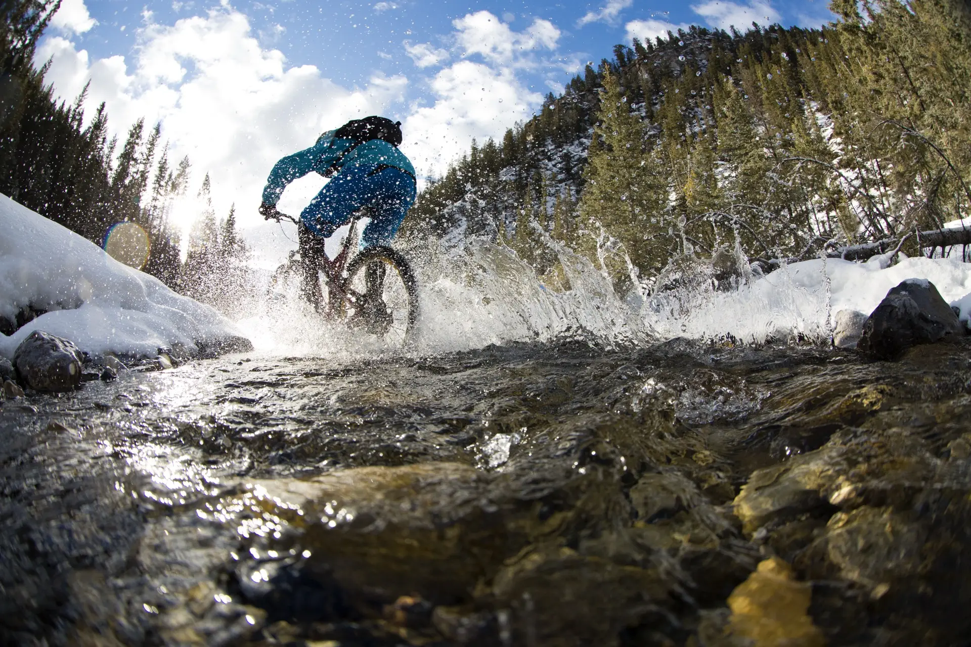 A mountain biker rides through a shallow rocky river plashing a wake behind.