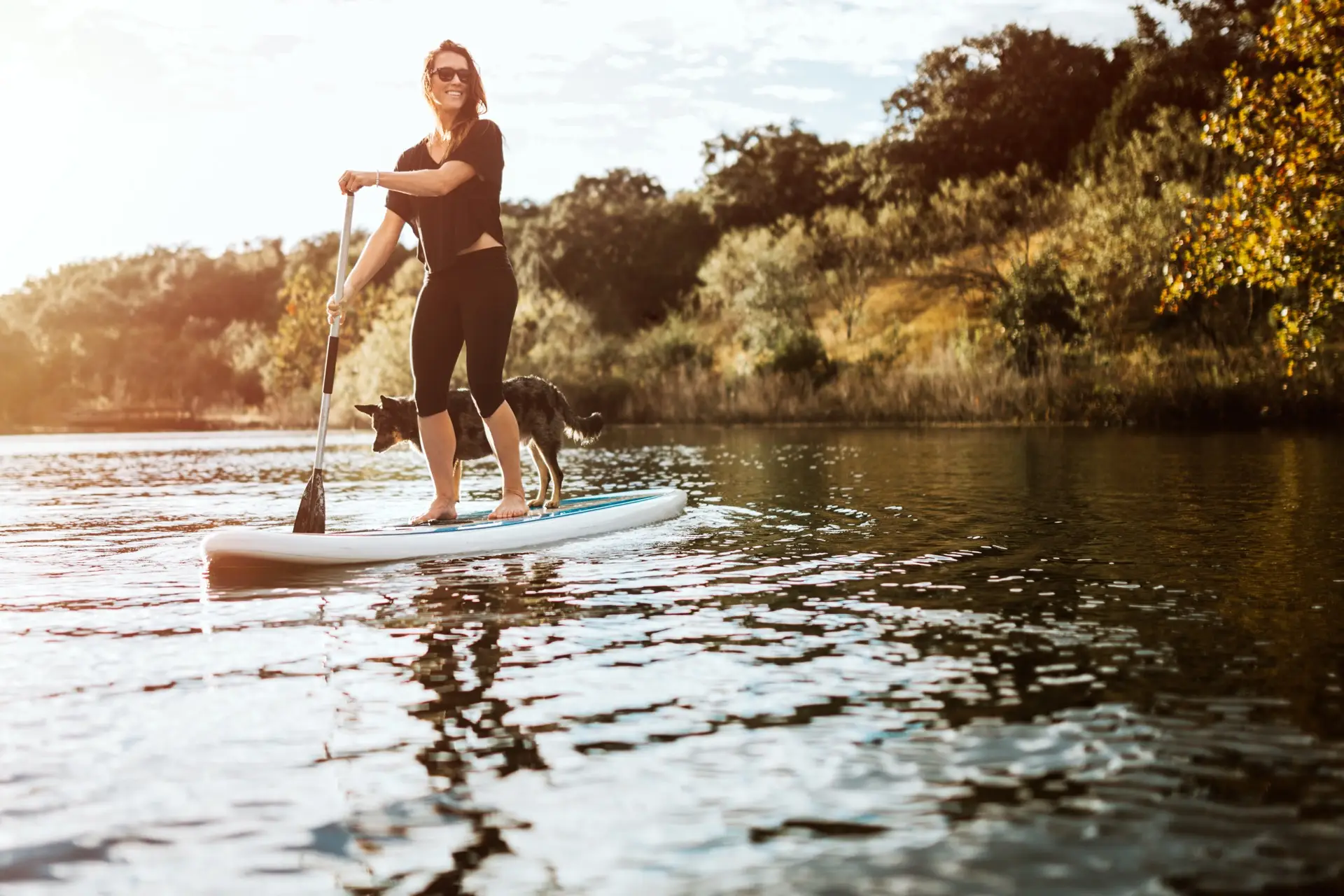 A young woman rides a standup paddleboard with her small black dog on a river.